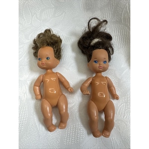 May include: Two vintage baby dolls with brown hair and blue eyes. The dolls are nude, with tan skin, and have articulated arms and legs. One doll has short, curly hair, while the other has long, wavy hair. The dolls are lying on a white patterned surface.