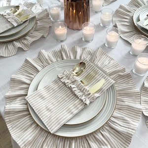 May include: A table setting with beige and white striped placemats, plates, and napkins. Gold-colored cutlery rests on the napkins. Small candles in glass holders and a copper vase with flowers complete the elegant dining arrangement.