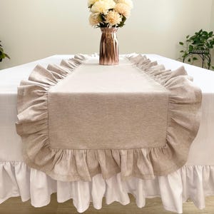 May include: A beige linen table runner with ruffled edges. The runner is centered on a white tablecloth with a white ruffled skirt. A copper vase with cream-colored flowers sits on the table.