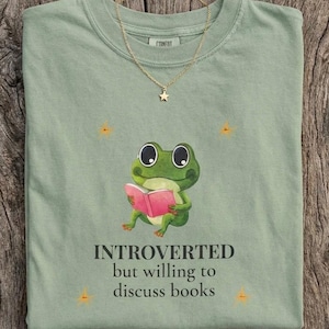 Puede incluir: Camiseta verde salvia con un dibujo de una rana de dibujos animados leyendo un libro rosa. El texto dice "INTROVERTED but willing to discuss books." Un collar dorado con un colgante de estrella está sobre la camiseta. La camiseta está sobre una superficie de madera.