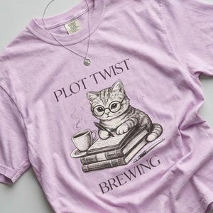 Puede incluir: Una camiseta de color lila claro con las palabras "PLOT TWIST BREWING" y un gráfico de un gato con gafas escribiendo en libros. Un collar plateado está sobre la camiseta.