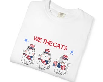 May include: White t-shirt featuring three cartoon cats wearing patriotic hats and bandanas, with the text "WE THE CATS" in red. The cats are holding American flags, and blue stars are scattered around the text.