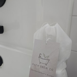 May include: A white aromatherapy bath tea bag with a tag that reads "BATH BREW CO. AROMATHERAPY BLEND." The tag features a bathtub illustration with a faucet and leaves. The bag is placed on the edge of a white bathtub.