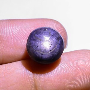 May include: A spherical, polished, purple gemstone displaying a star effect. The gem is held between fingers, highlighting its smooth surface and light refraction. The star pattern is clearly visible. A beautiful gem.