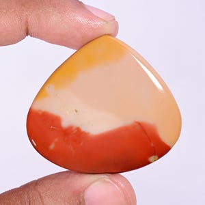 Puede incluir: Una piedra preciosa de jaspe Mookaite pulida en forma de lágrima con bandas de color horizontales. La piedra muestra una superficie lisa y brillante con una transición degradada desde un rojo terracota intenso en la base hasta crema y amarillo.
