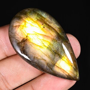 Può includere: Una pietra preziosa di labradorite a forma di goccia con una superficie lucida che presenta riflessi iridescenti in tonalità giallo brillante, oro e marrone scuro. La pietra mostra fratture interne naturali e una finitura liscia e riflettente.