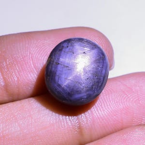 May include: A smooth, oval-shaped, purplish-blue gemstone. The gem has a polished surface with subtle internal lines and a slight sheen. The stone is held between fingers, highlighting its colour and texture. This gemstone could be used in jewellery.