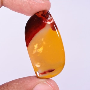 May include: A polished, teardrop-shaped cabochon gemstone featuring a smooth surface with a distinct color gradient. The stone displays a deep, dark red section at the top and bottom, transitioning into a vibrant, warm mustard yellow center.
