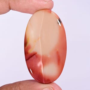May include: An oval-shaped cabochon gemstone featuring a smooth, polished surface with a blend of creamy beige, soft pink, and deep red color variations, including a small dark inclusion near the upper right edge of the stone.