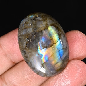 Puede incluir: Gema de labradorita de forma ovalada con superficie pulida. La piedra muestra una mezcla de tonos grises, marrones y azules, con destellos iridiscentes de luz amarilla, verde y azul. La gema se sostiene en una mano sobre un fondo negro.