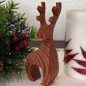 May include: A festive holiday arrangement featuring a wooden reindeer figurine, a small evergreen bush with red berries and pinecones, and a white candle decorated with red leaf accents. The items are arranged on a white tablecloth.