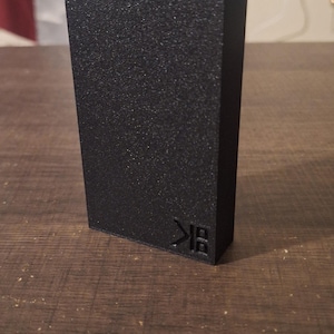 Puede incluir: Un objeto rectangular negro con una superficie texturizada. El objeto tiene un logotipo en la esquina inferior derecha. El objeto está sobre una superficie de madera.