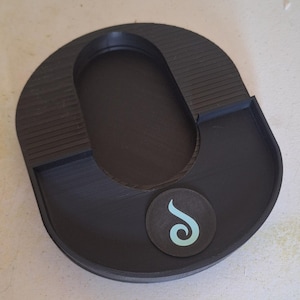 May include: A black plastic tray with a U-shaped design featuring a central oval cutout, ribbed side sections, and a circular emblem on the front surface displaying a light blue stylized swirl graphic. The tray has a smooth, matte finish.