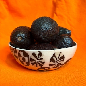 May include: A white ceramic bowl with a black and white floral pattern is filled with several dark, ripe avocados. The bowl sits on an orange surface, creating a vibrant contrast. The avocados have a rough, textured skin.
