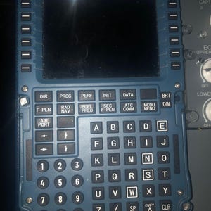 May include: A dark blue aircraft flight management system control unit featuring a rectangular screen, a numeric keypad, and an alphabetical keyboard with labeled buttons including DIR, PROG, PERF, INIT, DATA, BRT, DIM, F-PLN, RAD NAV, FUEL PRED, SEC F-PLN, ATC COMM, and MCDU MENU.