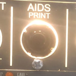 Peut inclure: Un panneau de contrôle noir avec un bouton rotatif blanc marqué FLOOD LT PED avec les réglages OFF et BRT. Deux interrupteurs à bouton-poussoir circulaires sont marqués AIDS PRINT et DFDR EVENT, tous deux éclairés par une lumière blanche vive.