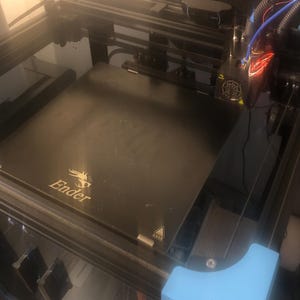 May include: A black 3D printer featuring a square build plate with the Ender logo printed in gold. The machine includes a black metal frame, a print head with visible wiring, a mounted webcam, and a light blue plastic corner component.
