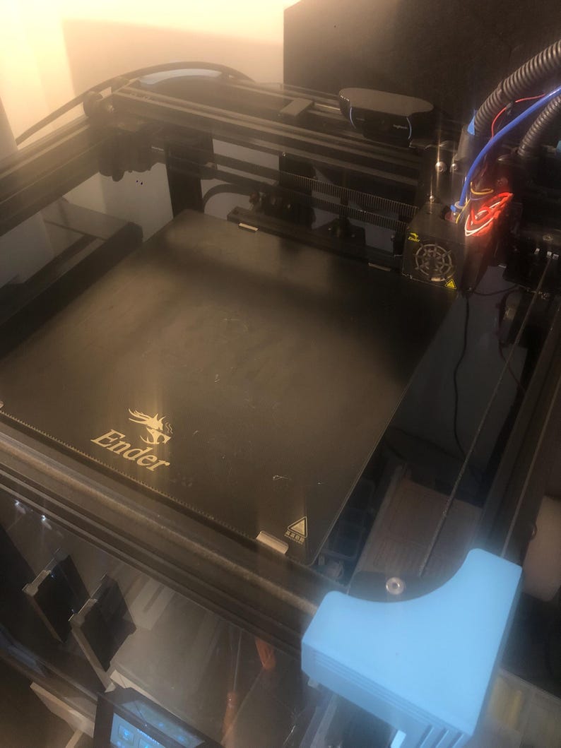 May include: A black 3D printer featuring a square build plate with the Ender logo printed in gold. The machine includes a black metal frame, a print head with visible wiring, a mounted webcam, and a light blue plastic corner component.
