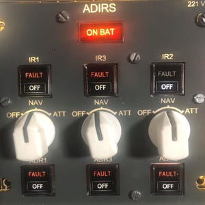May include: A dark gray ADIRS 221 VU control panel featuring three white rotary knobs labeled OFF, NAV, and ATT. The panel includes illuminated indicators labeled ON BAT, IR1, IR2, IR3, ADR1, ADR2, and ADR3, with FAULT and OFF text displayed on buttons.