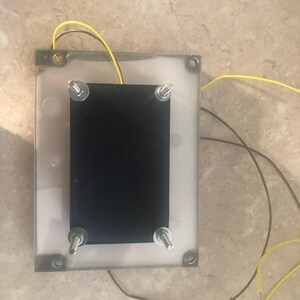 May include: A rectangular electronic component featuring a clear plastic base and a solid black rectangular plate secured by four metal bolts with nuts at each corner. Two thin yellow wires and one brown wire extend from the device.