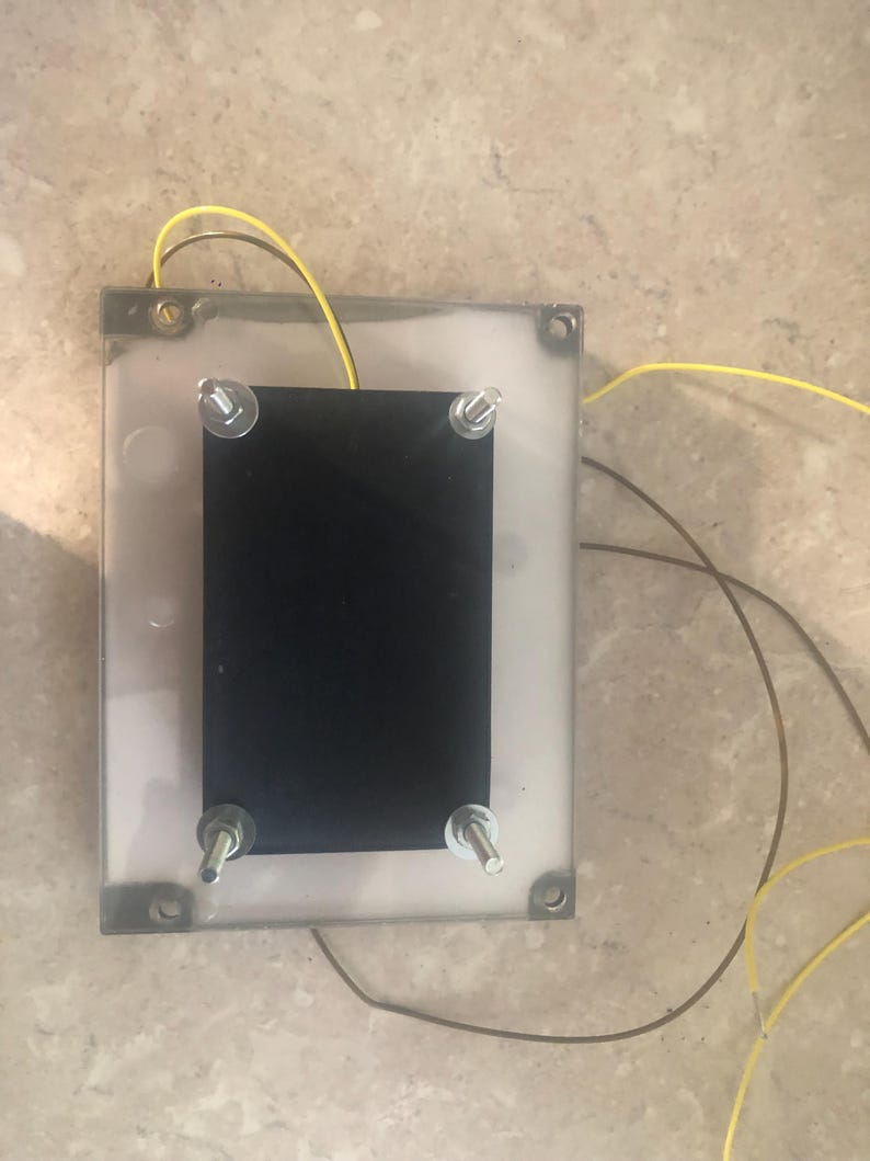 May include: A rectangular electronic component featuring a clear plastic base and a solid black rectangular plate secured by four metal bolts with nuts at each corner. Two thin yellow wires and one brown wire extend from the device.