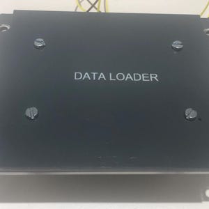 Peut inclure: Un appareil de chargement de données électronique noir et rectangulaire portant l'inscription DATA LOADER en blanc au centre. L'unité comporte quatre vis à tête plate fixant le panneau supérieur et des trous de montage aux coins avec des fils jaunes.