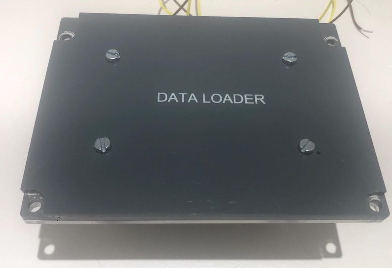 May include: A rectangular black electronic data loader device featuring the text DATA LOADER printed in white on the centre. The unit has four flat-head screws securing the top panel and mounting holes at each corner with thin yellow wires visible.
