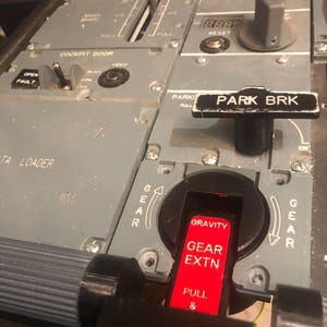 May include: A grey aircraft cockpit centre pedestal panel featuring a black parking brake lever labelled PARK BRK, a red gravity gear extension handle labelled GRAVITY GEAR EXTN PULL, a cockpit door control switch, and a speed brake lever.
