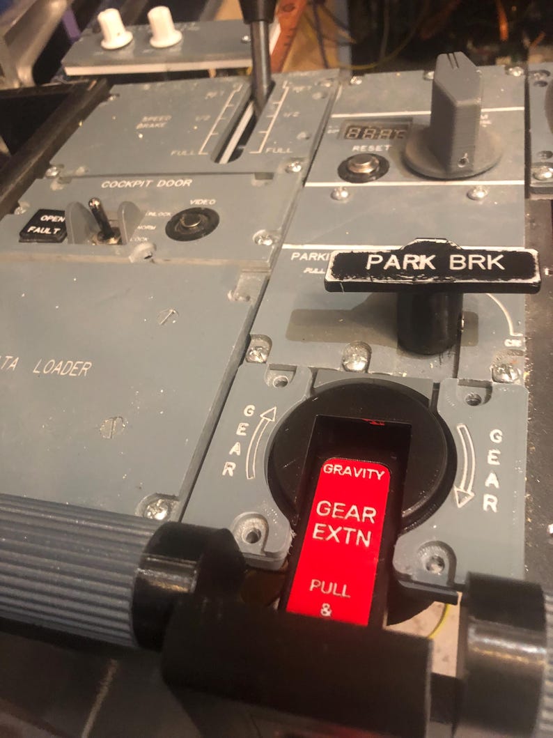 May include: A grey aircraft cockpit centre pedestal panel featuring a black parking brake lever labelled PARK BRK, a red gravity gear extension handle labelled GRAVITY GEAR EXTN PULL, a cockpit door control switch, and a speed brake lever.