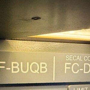 Peut inclure: Une plaque d'identification en métal gris avec le texte blanc F-BUQB et SECAL CODE FC-DG. La plaque est fixée par deux vis de couleur argentée sur les côtés gauche et droit, placée sous une source de lumière.