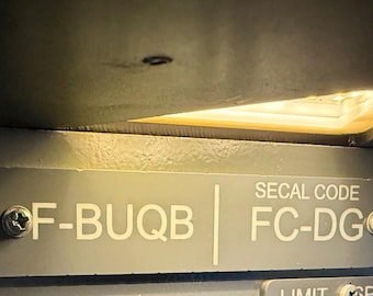 May include: A grey metal identification plate featuring white text that reads F-BUQB and SECAL CODE FC-DG. The plate is secured with two silver-coloured screws on the left and right sides, positioned under a light source.