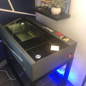 May include: A dark grey laser cutter machine with a large black glass lid and a glowing circular power button on the top panel. The device features a red emergency stop button on the side and is mounted on a black metal stand with blue LED lighting.