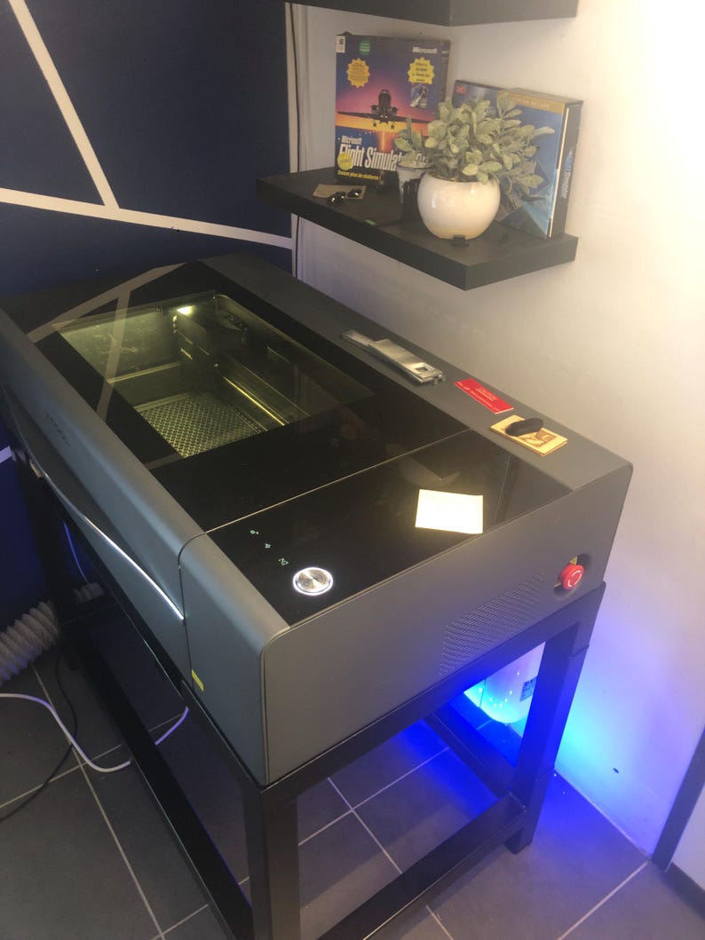 May include: A dark grey laser cutter machine with a large black glass lid and a glowing circular power button on the top panel. The device features a red emergency stop button on the side and is mounted on a black metal stand with blue LED lighting.