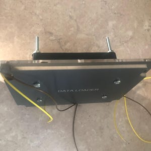 May include: A rectangular grey electronic component labelled DATA LOADER with a black base, a clear acrylic top layer, and two vertical metal bolts. Thin brown and yellow wires extend from the sides of the device, which is secured by four screws.