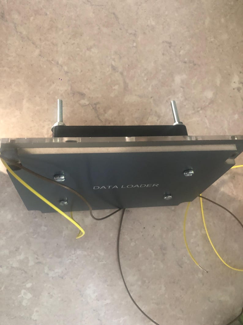 May include: A rectangular grey electronic component labelled DATA LOADER with a black base, a clear acrylic top layer, and two vertical metal bolts. Thin brown and yellow wires extend from the sides of the device, which is secured by four screws.
