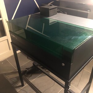 May include: A black xTool laser cutter machine featuring a rectangular, translucent green lid and a matte black metal frame stand. The device has a sleek, industrial design with a prominent xTool logo on the front panel and a small black control box.