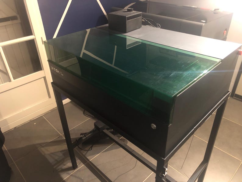 May include: A black xTool laser cutter machine featuring a rectangular, translucent green lid and a matte black metal frame stand. The device has a sleek, industrial design with a prominent xTool logo on the front panel and a small black control box.