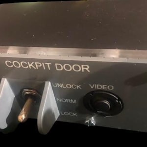 May include: A gray rectangular cockpit door control panel featuring a metal toggle switch protected by two vertical guards, a black circular push button labeled VIDEO, and white text reading COCKPIT DOOR, UNLOCK, NORM, and LOCK, with 119VU printed on the side.
