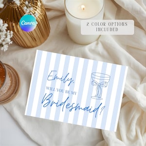 Puede incluir: Una tarjeta a rayas azules y blancas con el texto "Emily, Will You Be My Bridesmaid?" y un dibujo de una copa de champán. La tarjeta está sobre una tela blanca, con una vela y un jarrón dorado en el fondo. También es visible el texto "2 COLOR OPTIONS INCLUDED".