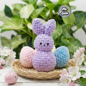 Puede incluir: Un conejito de ganchillo lavanda hecho a mano con una cesta de huevos de Pascua de colores pastel. El conejito tiene orejas largas y un cuerpo redondo. Los huevos son rosas, azules y amarillos. La escena está decorada con flores blancas y follaje verde.