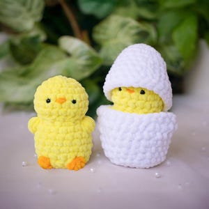 Könnte beinhalten: Zwei handgefertigte Häkelküken, eines stehend und eines in einem weißen Ei. Das stehende Küken ist gelb mit orangefarbenen Füßen und einem kleinen orangen Schnabel. Das Küken im Ei ist ebenfalls gelb, mit einer weißen Eierschale.