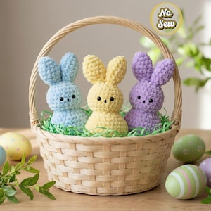 Könnte beinhalten: Ein geflochtener Korb mit drei gehäkelten Osterhasen in Pastellblau, Gelb und Lila, eingebettet in grünes Kunstgras. Dekorierte Ostereier in verschiedenen Farben umgeben den Korb und schaffen eine festliche Frühlingsszene.