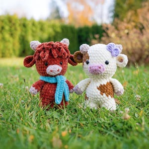 May include: Two handmade crochet cow plushies stand in green grass. One is red-brown with a blue scarf, the other is white with brown spots and a purple flower. They have small horns and are holding hands.