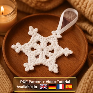 May include: A white, crocheted snowflake ornament with a ribbon for hanging, resting on a wooden plate. A lit candle in a glass jar and a knitted blanket are in the background. The image includes text: "PDF Pattern + Video Tutorial Available in".
