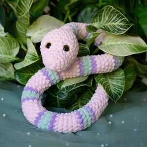 Könnte beinhalten: Ein handgefertigtes Schlangen-Spielzeug aus Plüsch in Rosa, Lila und Grün. Die Schlange hat schwarze Augen und ist vor grünen Blättern aufgerollt. Das Spielzeug ist aus weichem, strukturiertem Garn gefertigt.