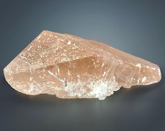 May include: A large, rough-hewn crystal with a peach hue. The crystal has a flat, angled top and a jagged, uneven base. The surface has a textured appearance with areas of white and light brown.