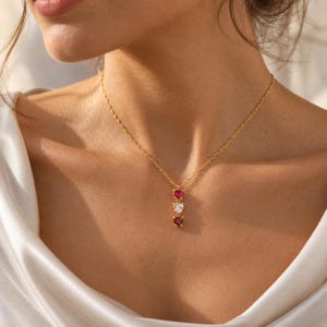 May include: A gold chain necklace featuring a vertical pendant with three small heart-shaped gemstones arranged in a row. The top stone is bright red, the middle stone is clear white, and the bottom stone is a deep dark red.