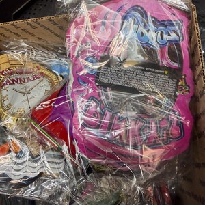 May include: A box filled with various items wrapped in clear plastic. A pink package with the word "Bratz" in a stylized font is visible. Other items include a clock-shaped item with the word "ANNAB" and a red object.