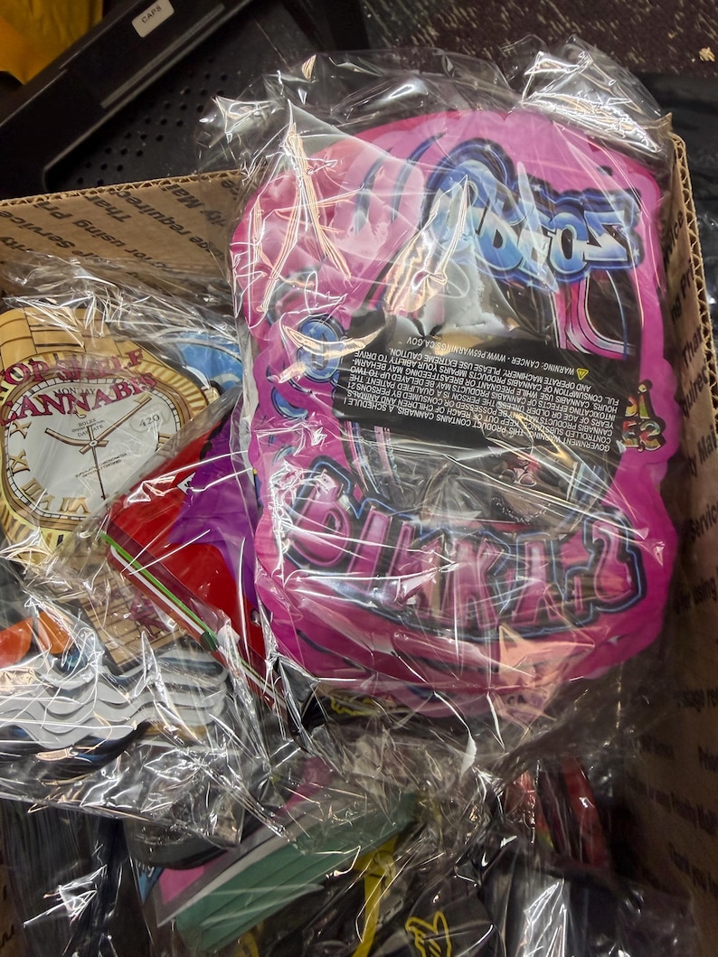 May include: A box filled with various items wrapped in clear plastic. A pink package with the word "Bratz" in a stylized font is visible. Other items include a clock-shaped item with the word "ANNAB" and a red object.