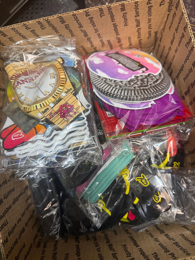 May include: A cardboard box filled with various stickers and accessories, including clock-themed designs, colorful circular stickers, and black accessories. The items are packaged in clear plastic bags, ready for shipping.